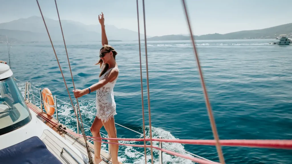 A woman in a white dress enjoying the breeze on a sailboat with the ocean in the background.