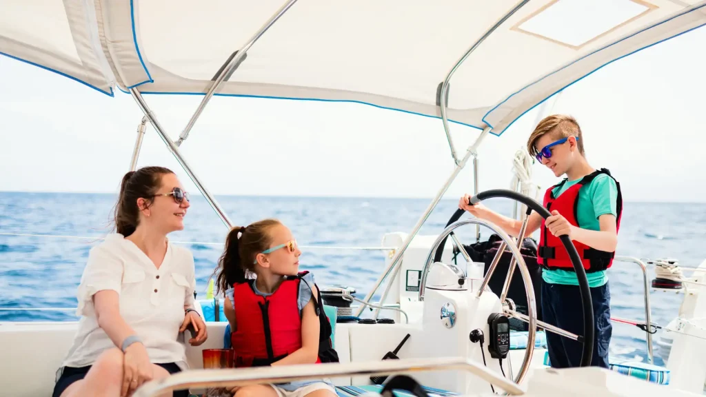 Three people on a sailboat, two wearing life jackets, while one steers the boat on a sunny day at sea