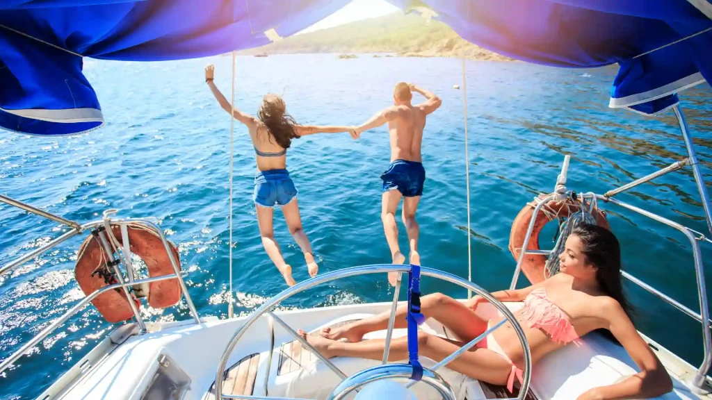A couple jumping off a boat into the water, holding hands while others relax on the boat.